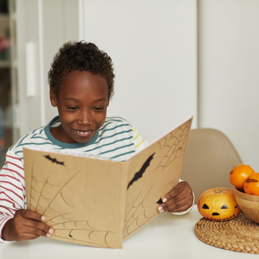 School-aged child sitting at a table and holding and reading from a Halloween-inspired storybook.