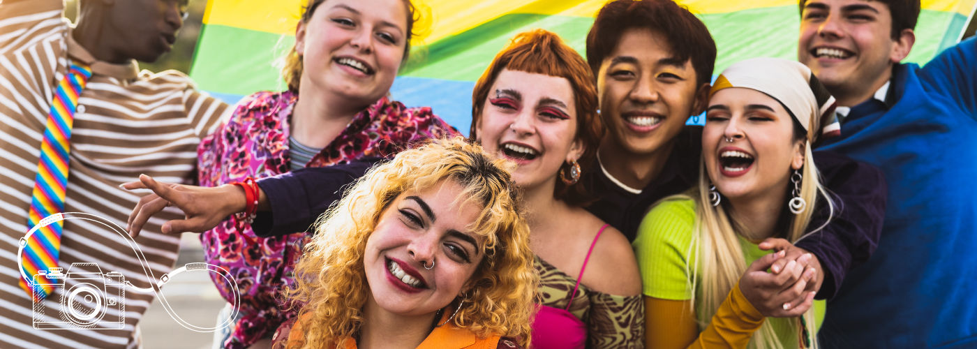 Six smiling and colourfully-dressed individuals celebrating Pride and holding a rainbow flag behind their heads.