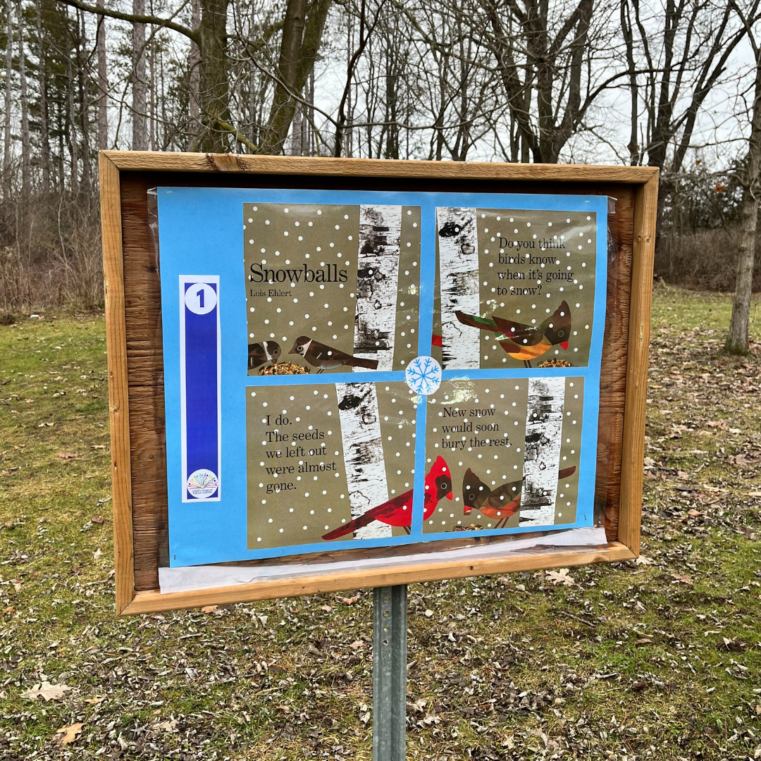 A Storybook Walk storyboard. The storyboard is located along a tree-covered trail in a forest or park-like setting.