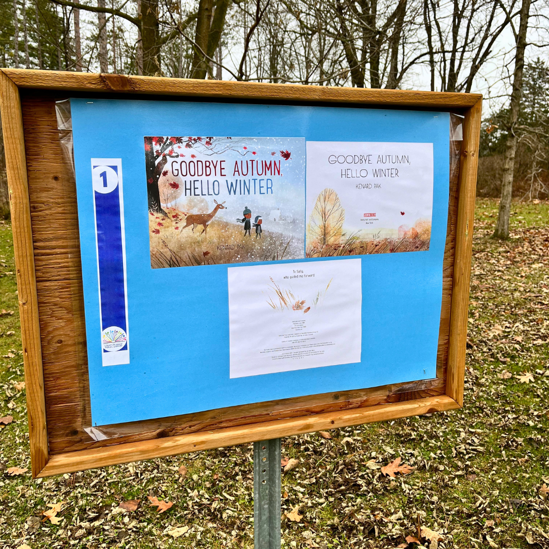 A Storybook Walk storyboard. The storyboard is located along a tree-covered trail in a forest or park-like setting.